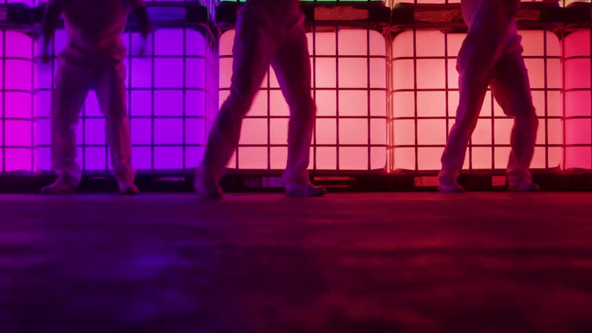 A lowangle shot captures the legs and feet of three dancers moving rhythmically against a colorful neon backdrop made of large stacked illuminated grid squares the scene is highly stylized with vibrant pink and purple lighting that casts dramatic sha