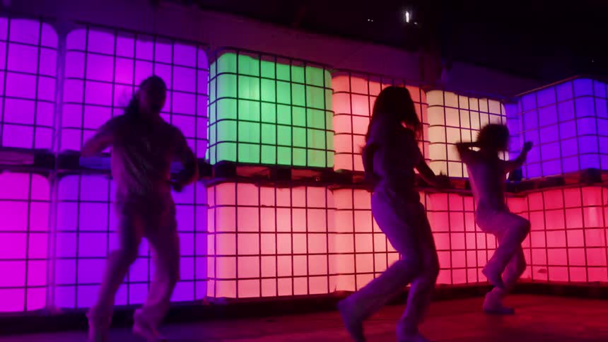 Three dancers are shown in silhouette against a brightly lit wall composed of stacked translucent containers illuminated by shifting neon colors like pink red and green the overall scene has a dark stylized club or rave atmosphere with the dancers in