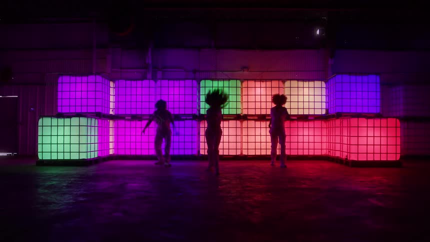 Three women dance in front of a stack of colorful illuminated cubes