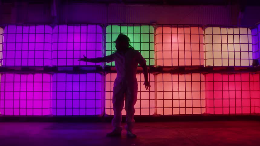 A person in a white jumpsuit performs dynamic dance movements in a warehouse or studio against a wall of stacked plastic totes brightly lit with shifting multicolored led lights the colors transition dramatically between purple green orange and whit