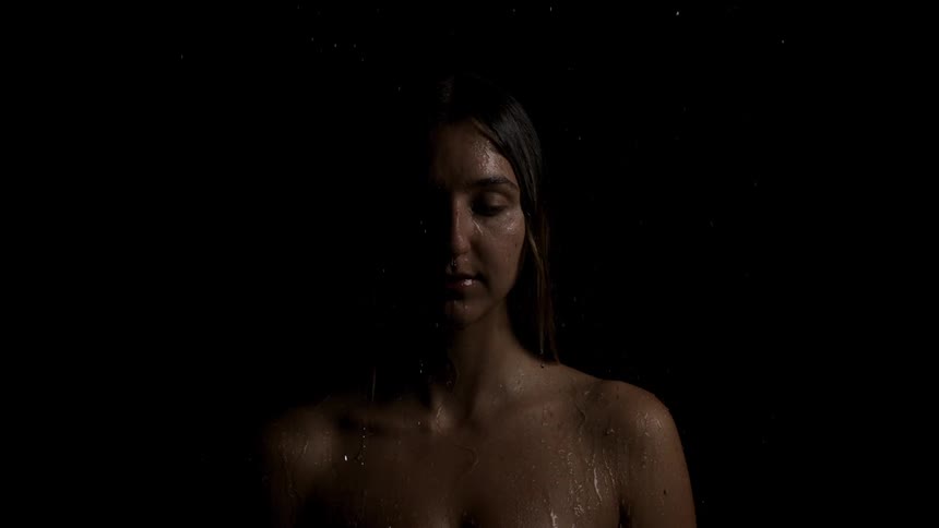 A closeup shot focuses on a womans shoulders and face in neartotal darkness with one side dramatically illuminated by a dim concentrated light source water droplets continuously fall on her skin suggesting a scene in a shower or under rain evoking