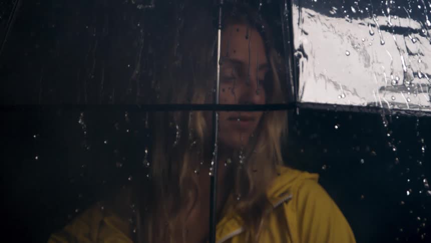 Woman in a yellow raincoat watches the rain through a transparent umbrella