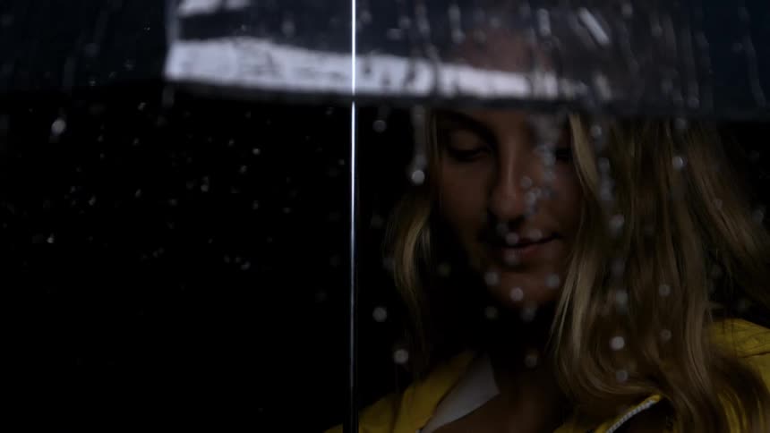 A woman in a yellow rain jacket smiles slightly and looks offcamera while holding a clear umbrella as rain falls heavily around her the shot is dark and moody with the focus on the rainstreaked umbrella and the womans face