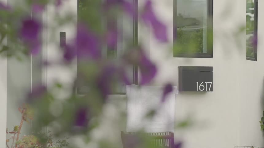 Through a blur of purple flowers a modern white house faade comes into focus with blackframed windows a wallmounted mailbox labeled 1617 and a chair draped with a light towel