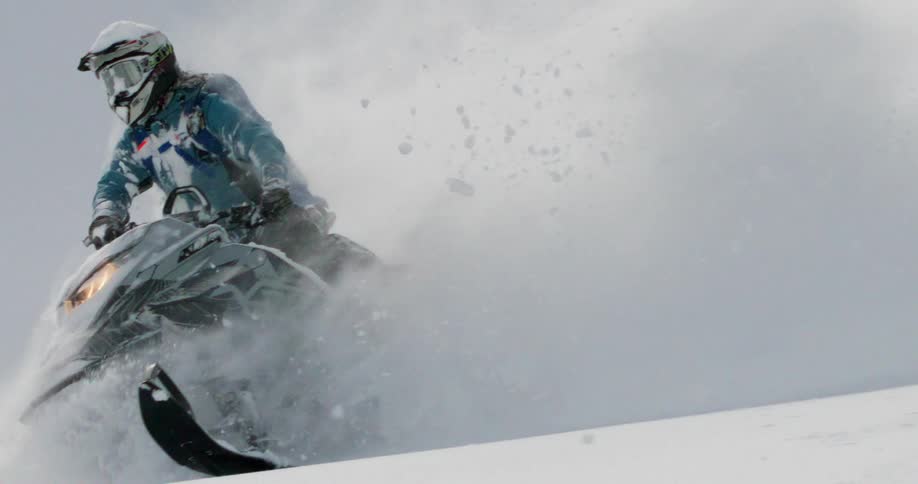 A person wearing a helmet and blue jacket is riding a snowmobile at high speed up a steep slope covered in deep fresh powder the snowmobile kicks up a massive plume of white snow momentarily obscuring the rider and creating a dynamic powerful image