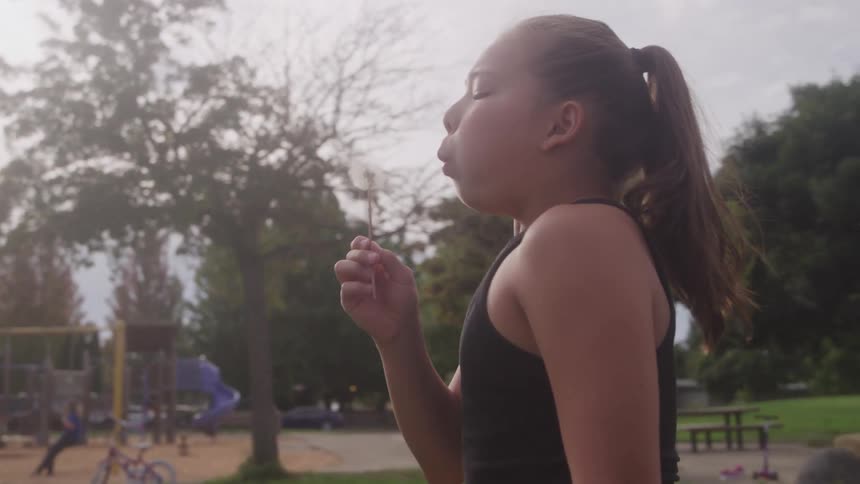 A young girl in a park blows a dandelion seed head cheeks puffed and expression focused with a playground in the background