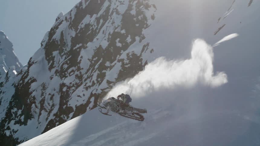A person wearing a helmet and winter gear rides a snowmobile at speed across a large open snowy slope kicking up a substantial cloud of powder the snowmobile appears to be moving laterally across the steep face of the snowcovered mountain with a brig