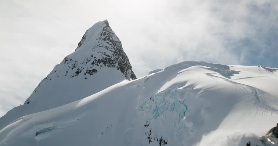 The video clip shows an extreme long shot of a majestic snowcovered mountain peak that sharply juts out against a bright sky the foreground reveals a steep slope of snow and ice with visible ski or snowboard tracks curving down the ridge