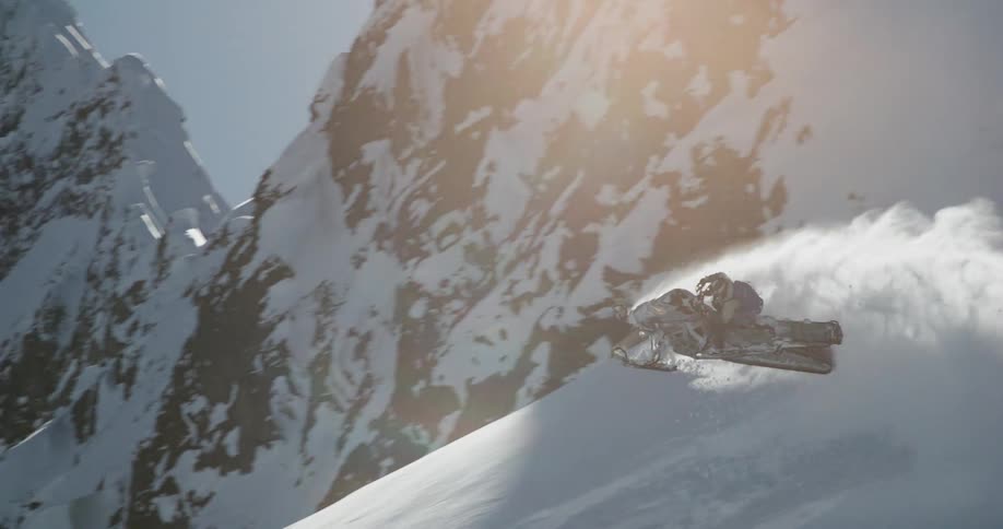 A person wearing a dark helmet and gear rapidly rides a snowmobile up a very steep slope of deep snow kicking up a large blinding white cloud of powder the snowmobile maneuvers quickly and sharply with the massive mountain face visible in the backgrou