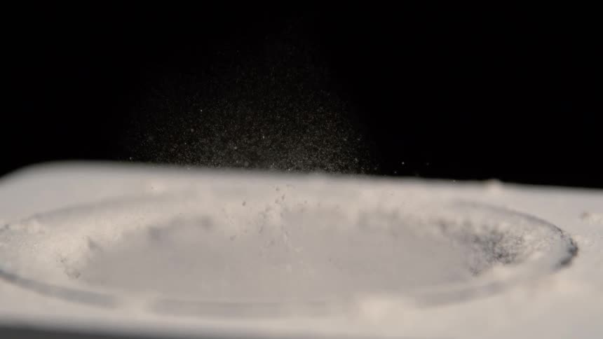 A burst of fine white powder dances above a vibrating circular surface against a black background