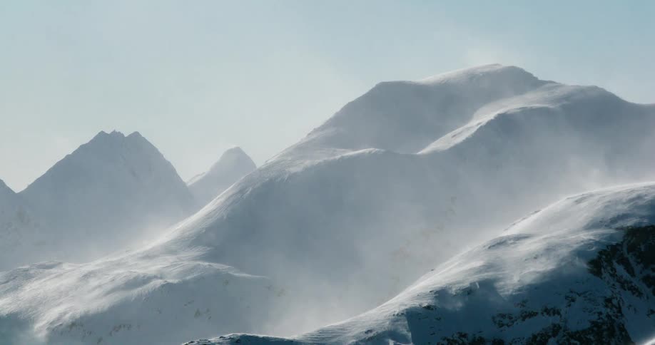 A slow breeze causes powdered snow to off snow covered mountains below a mostly cloudless sunny sky
