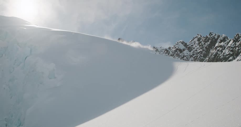 This video is a dramatic wide shot of a snowboarder or skier descending a steep fresh powder snow slope on a bright sunny day the rider carves a turn kicking up a large billowing cloud of powder snow as they move across the pristine white mountain f