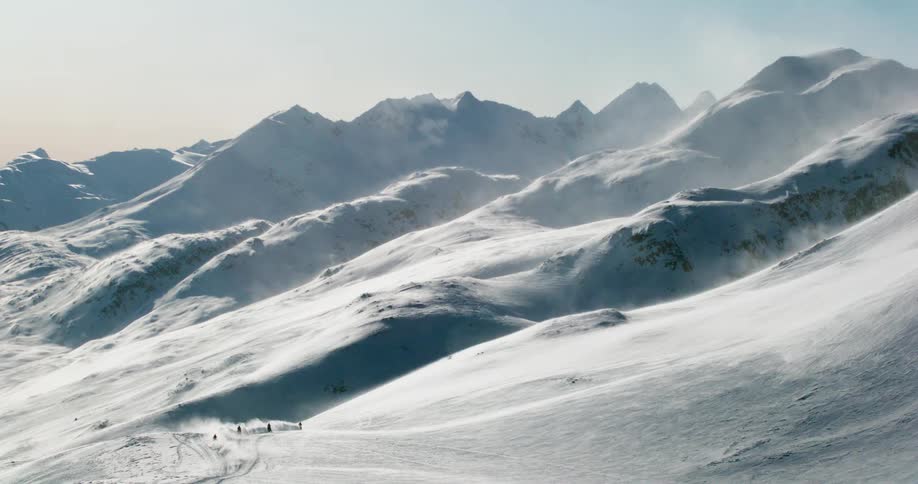 People ride snowmobiles across mountainous terrain creating a cloud of snow on a winter day