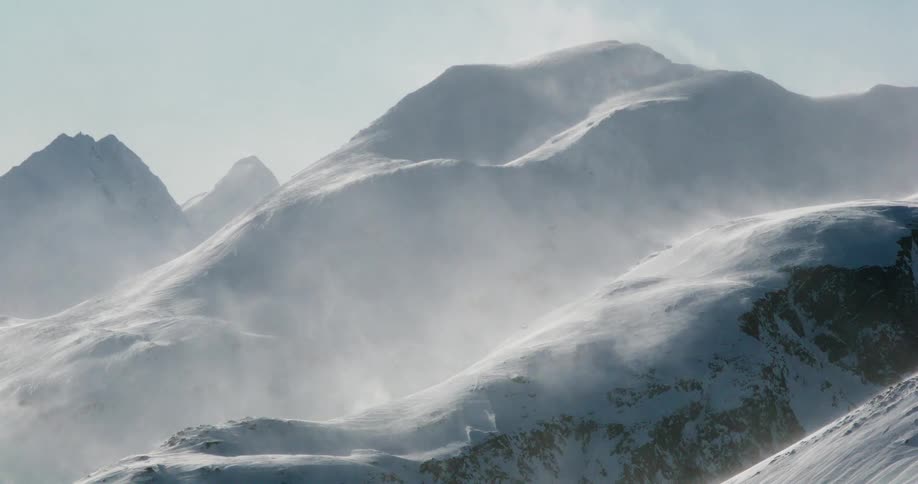 Windwhipped spindrift lifts off sunlit snowy ridgelines tracing the contours of rugged alpine peaks under a clear sky
