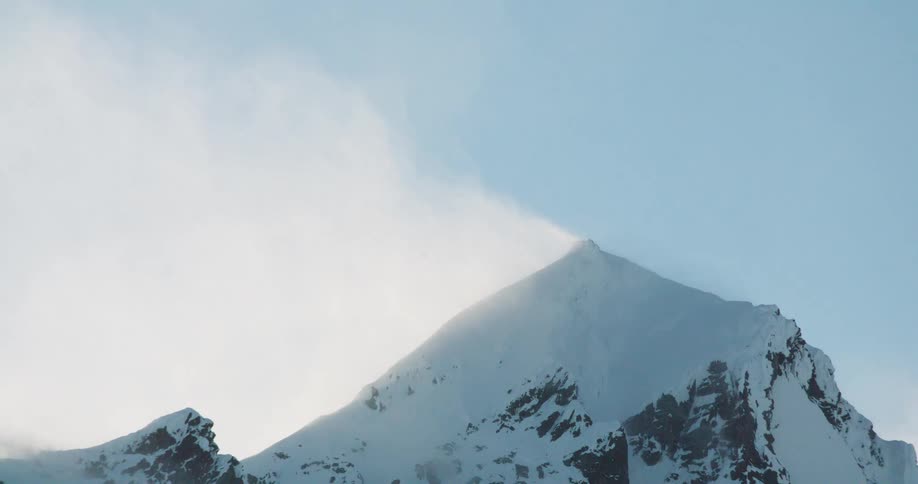 A snowcovered mountain peak fills the center of the frame with snow or vapor being blown off its summit under a light blue sky framed by the dark rocky mass of the mountain rising from the bottom