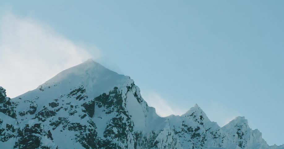 Wind blows the snow off the mountain peaks into the air on a winter day