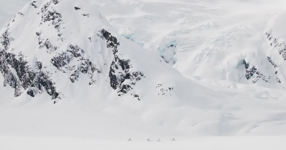 A small group trace a line across a vast white glacier on some type of vehicle beneath towering craggy cliffs streaked with blue ice