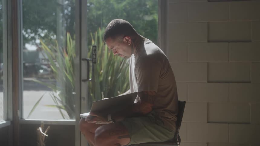 The video clip shows an indoor mediumclose shot of a man with tattoos on his arms and legs sitting in a chair next to a large window he is wearing a tshirt and shorts and is looking down intently at a dark notebook or binder resting on his lap
