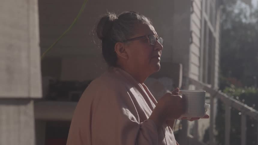 An elderly woman sits peacefully on her front porch at midday wearing glasses and holding a steaming cup of hot coffee as sunlight warms the quiet scene