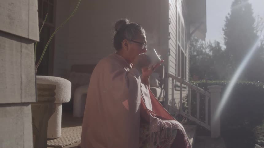 A woman wrapped in a shawl sips from a cup on a sunlit porch morning light streaming across the steps and garden