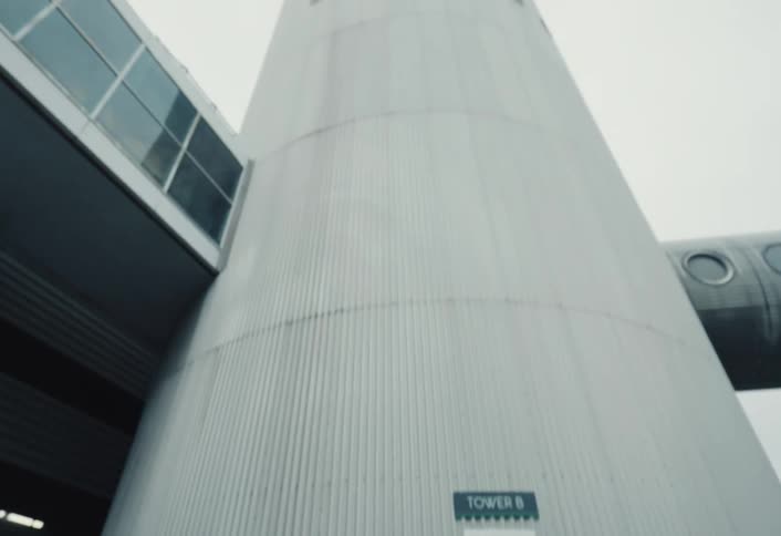 Person wearing a green jacket and black hat looks upward near a gray industrial building