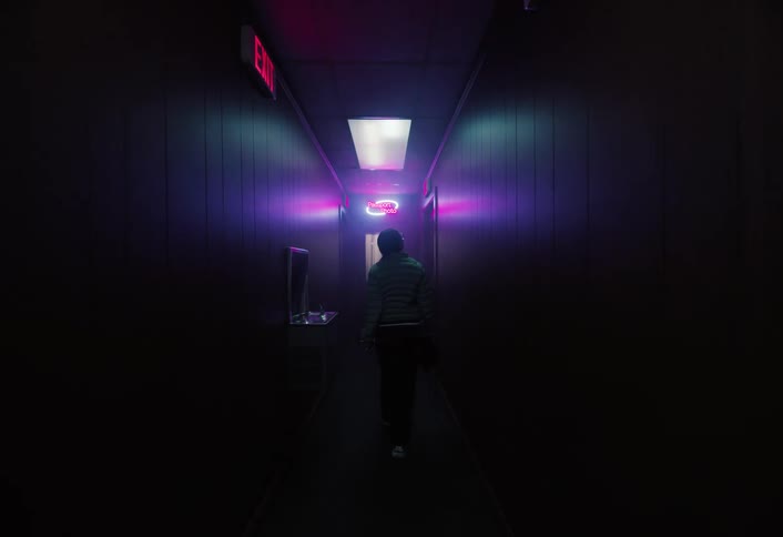 A person walks down a long dark hallway with woodpaneled walls toward the camera illuminated only by a single overhead fluorescent light and bursts of red and purple neon from the sides the scene has a dramatic mysterious or retro atmosphere remini