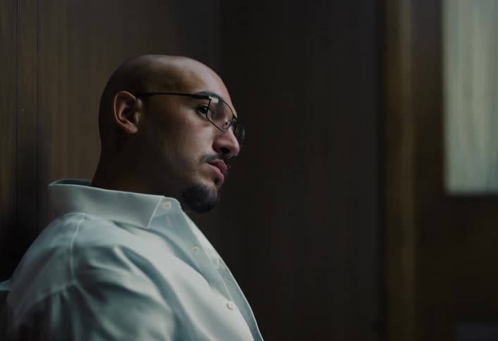 The video clip shows a closeup side profile of a man with a shaved head and beard wearing a white collared shirt and glasses he is seated against a dark background looking downward with a thoughtful or troubled expression
