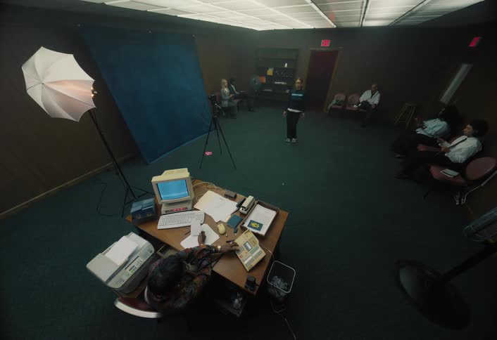 In a dim retro officephoto studio a person works at a cluttered desk with an old computer and rotary phones while a blue backdrop and umbrella light face a row of people waiting along the walls
