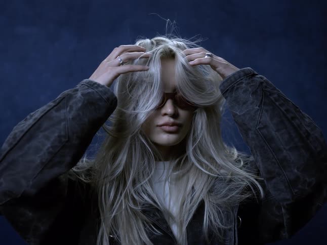The video clip is a closeup midshot of a young woman with long light blonde hair she is wearing sunglasses and a denim jacket and runs her hands through her hair against a dark blue background