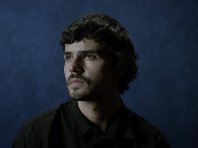 The video clip is a closeup midshot of a man with dark curly hair and a beard looking offcamera against a dark blue background he is wearing a dark collared shirt and has a thoughtful or serious expression on his face