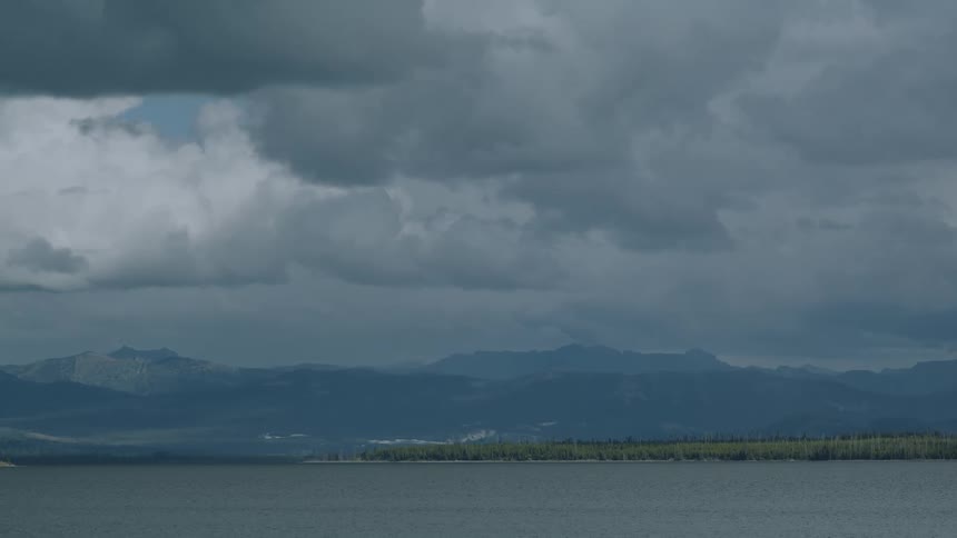 Dark heavy clouds fill the sky above a vast lake with a distant treelined shore and a mountain range visible on the horizon