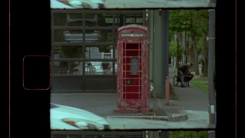 Car stops at an intersection near a red phone booth while a woman leans over a baby carriage near a car repair garage