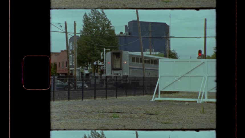 This vintage film clip shows a single train car slowly moving from right to left past a low fence in an urban industrial area the frame includes film sprockets and borders capturing buildings and a red signal light in the background
