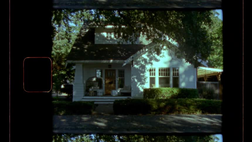 This vintage film clip is a slow zoomout shot of a charming traditional whitesided house with a porch and prominent multipaned windows the house is partially shaded by dense overhead trees and surrounded by neatly trimmed hedges