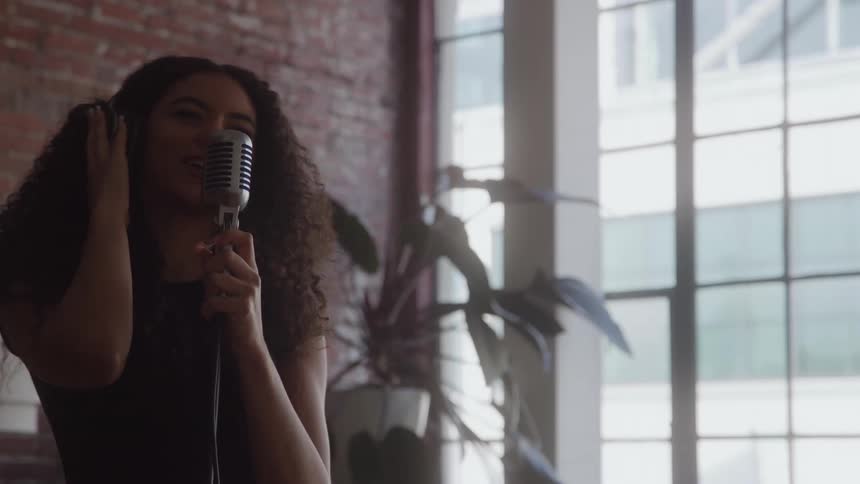 This is a mediumcloseup shot of a woman with long curly hair passionately singing into a vintage microphone with one hand touching her ear the setting appears to be a brightly lit loft or studio with an exposed brick wall and tall windows