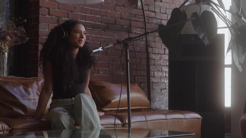 A young woman sits on a couch while talking into a microphone on a stand
