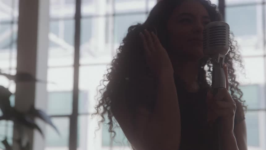 The face of a young woman with curly hair who has her hand on her ear while singing into the microphone