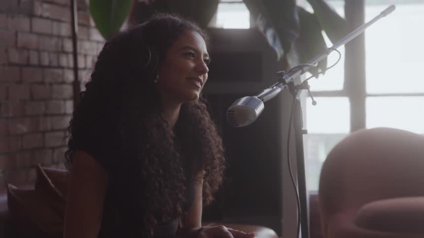This is a medium shot of a woman with long curly hair wearing headphones who is speaking into a large microphone on a stand she is sitting in a sunlit studio with an exposed brick wall using hand gestures as she talks