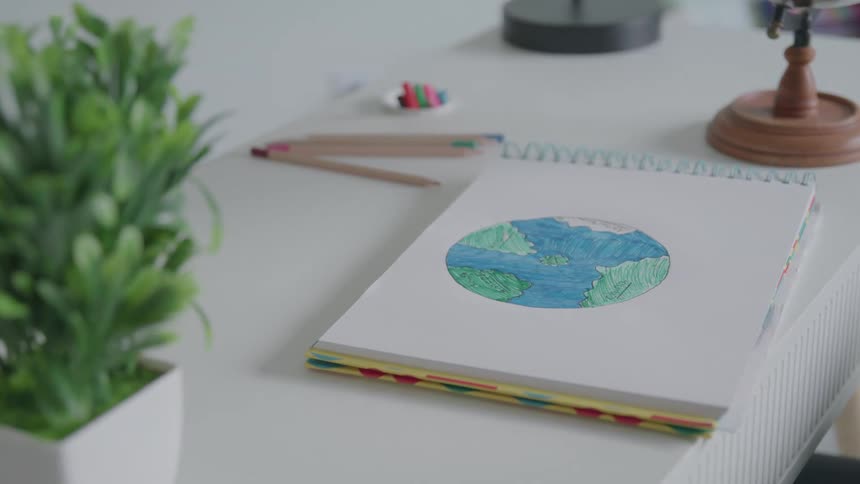 This is a static closeup shot of a white desk with a sketchpad open to a childs colorful drawing of the earth an artificial green plant is visible in the foreground with pencils and a small globe stand in the background
