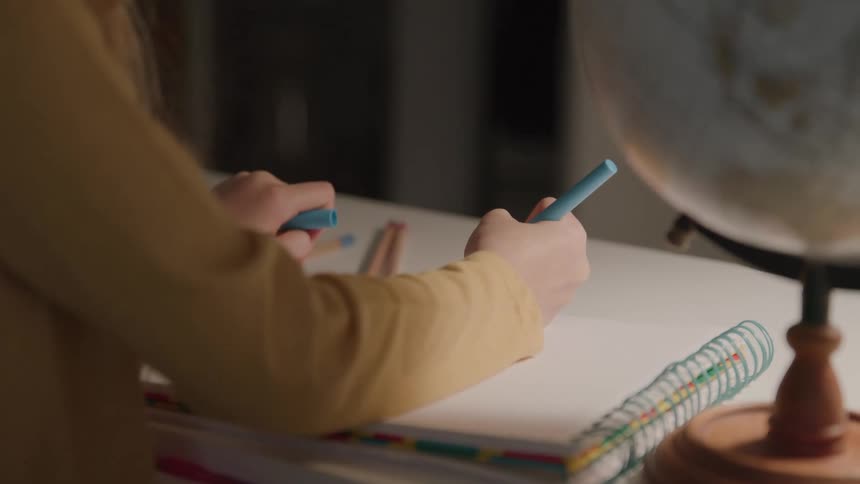 This is a closeup static shot focusing on the hands of a person in a mustardyellow longsleeve shirt writing or drawing with a blue marker in a notebook a large vintage globe is visible on the right side of the frame