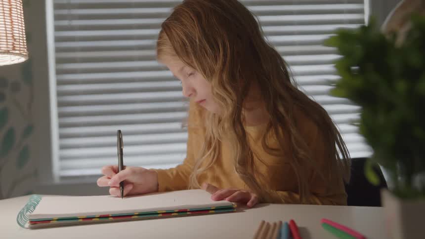 This is a closeup static shot of a young girl with long hair intently writing or drawing in a spiral notebook with a dark pen or pencil the scene is illuminated by a lamp on the left with the light filtering through window blinds in the background