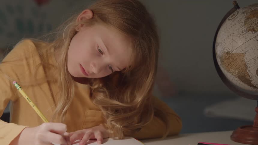 This is a closeup static shot of a young girl with long blonde hair concentrating intently as she writes with a pencil on a piece of paper a globe is visible on the desk to the right and the scene is dimly lit by a nearby light source
