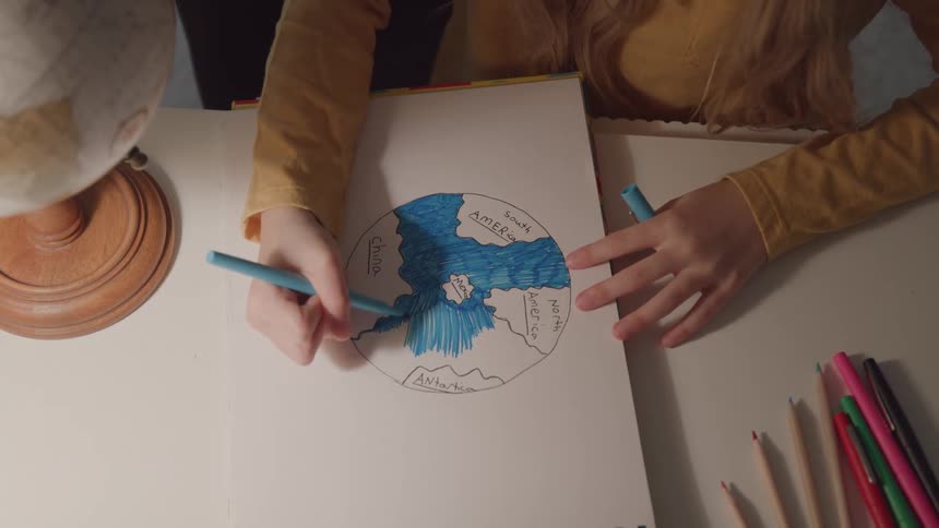 This is a static overhead shot of a persons hands coloring a handdrawn map of the world in a sketchpad specifically filling in the oceans with a blue marker a woodenbased globe is visible in the foreground on the left