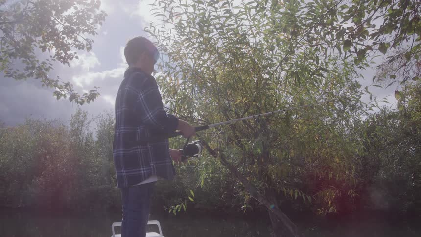 A boy wearing a plaid shirt is standing fishing in a lake with several trees around