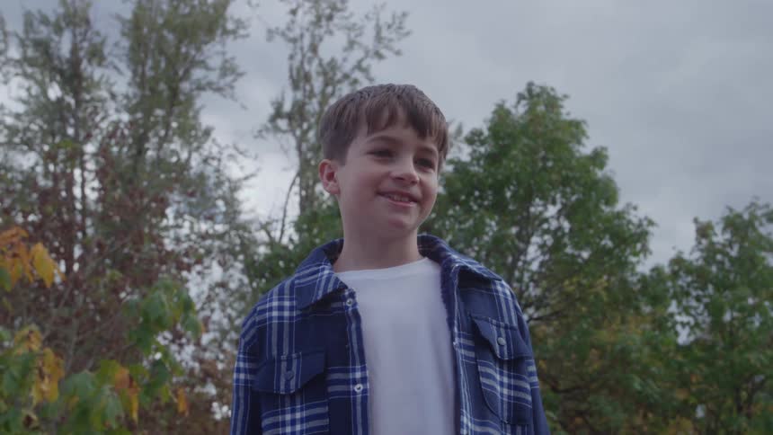 The video is a mediumcloseup shot of a young boy in a blue plaid shirt looking up and smiling in an outdoor setting he is surrounded by the green and yellow foliage of trees against a bright overcast sky