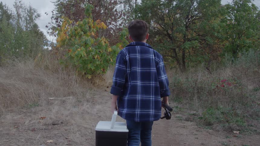 This is a static shot from behind of a young boy walking down a dirt path in a wooded area wearing a blue plaid shirt he is carrying a cooler in his right hand and what appears to be a fishing rod in his left