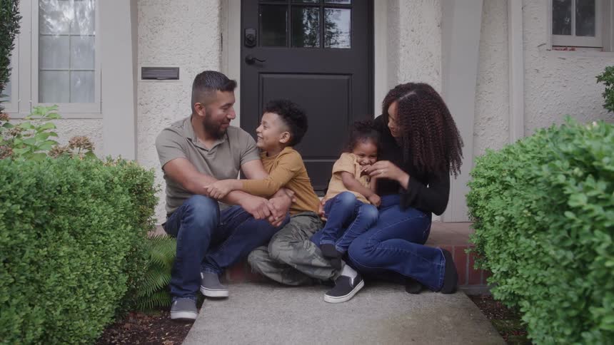 A mother and father sit in front of their home with their children