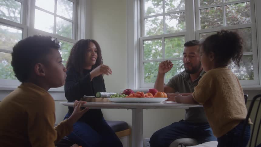 This is a mediumwide shot of a family enjoying a healthy snack together around a bright table next to tall multipaned windows the table is filled with fresh items like sliced cucumbers carrots and a variety of fruit