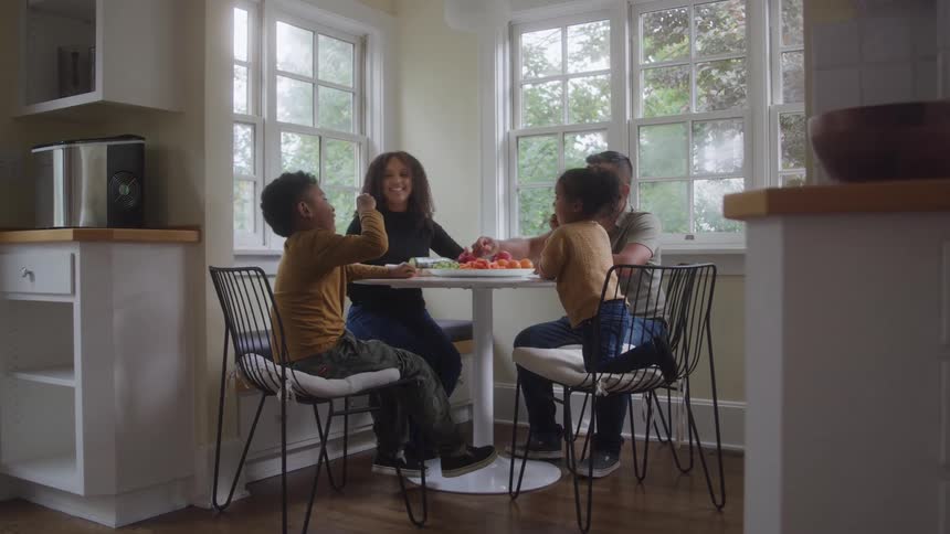The video is a wide shot of a family of four sitting around a small table in a bright modern kitchen and enjoying a snack together the scene is dominated by a large window with natural light and features a plate piled with fresh fruits and vegetables in