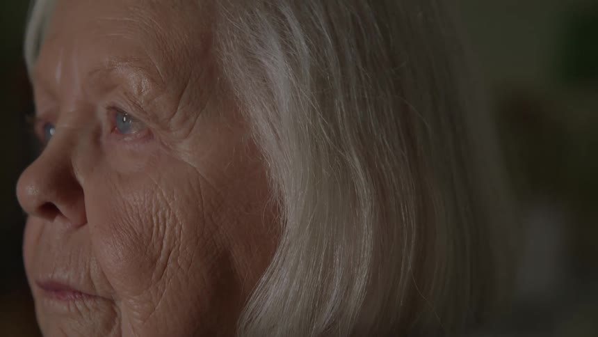 This is an extremely tight closeup profile shot focusing on the face of an elderly woman with white hair showing fine detail in her features and light blue eyes the shot has a shallow depth of field and a serious or contemplative tone